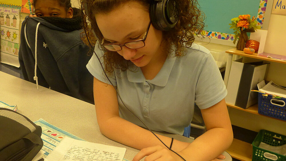 A youth wearing headphones interacting with a portable recorder on a desk in a classroom