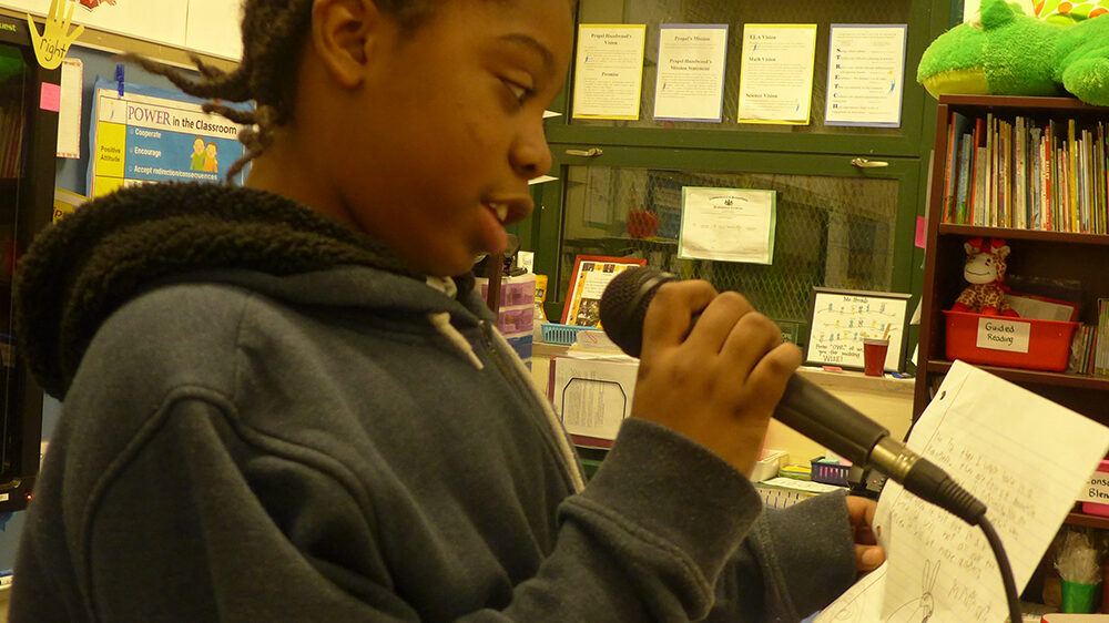 A youth reading a piece of paper and holding a microphone in a classroom