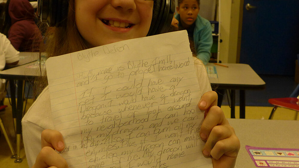 A youth wearing headphones holding a piece of notebook paper up to the camera in a classroom