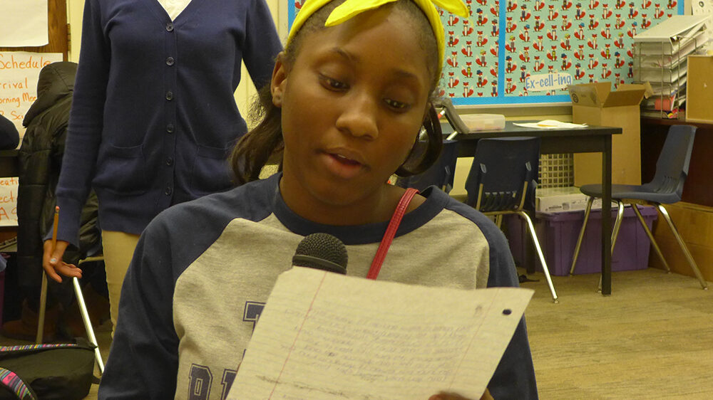 A youth reading a piece of paper holding a microphone in a classroom