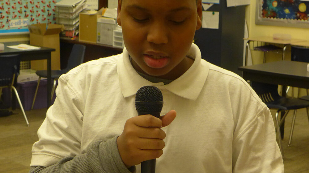 A youth holding a piece of paper speaking into a microphone in a classroom