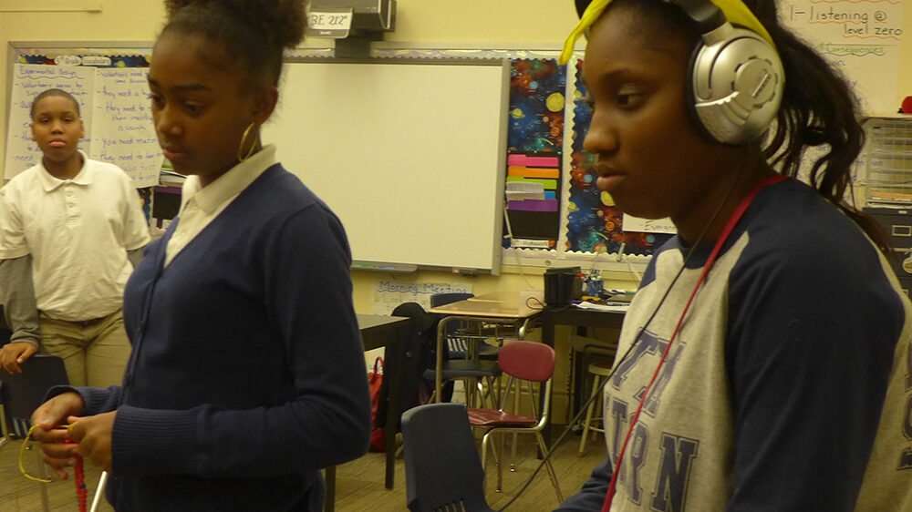 Two youth in a classroom, one standing, one sitting while wearing headphones and holding a microphone