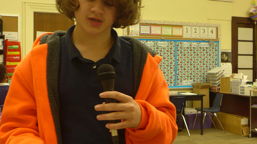 A youth holding a microphone reading a piece of paper in a classroom