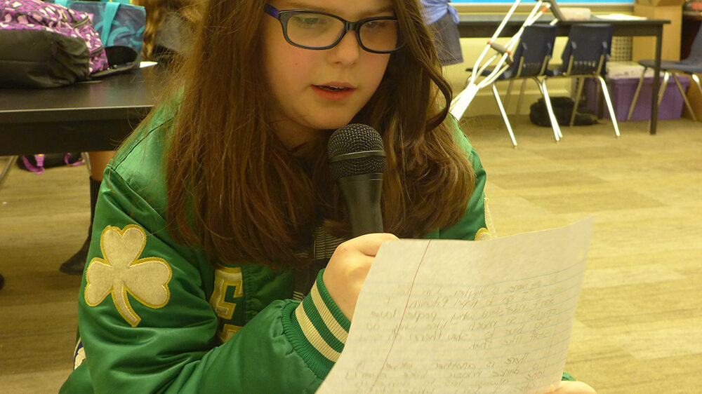 A youth holding a piece of paper reading into a microphone in a classroom