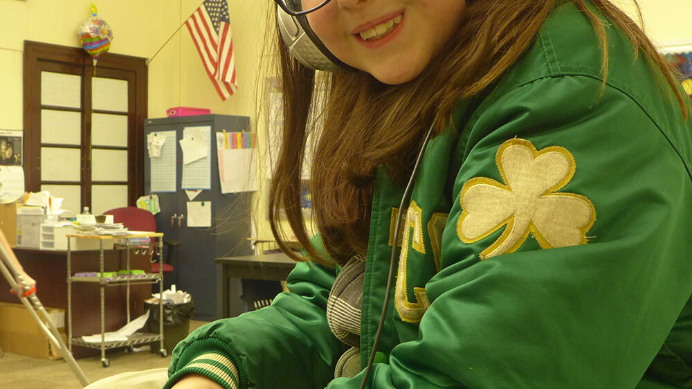 A youth wearing headphones holding a microphone smiling at the camera in a classroom