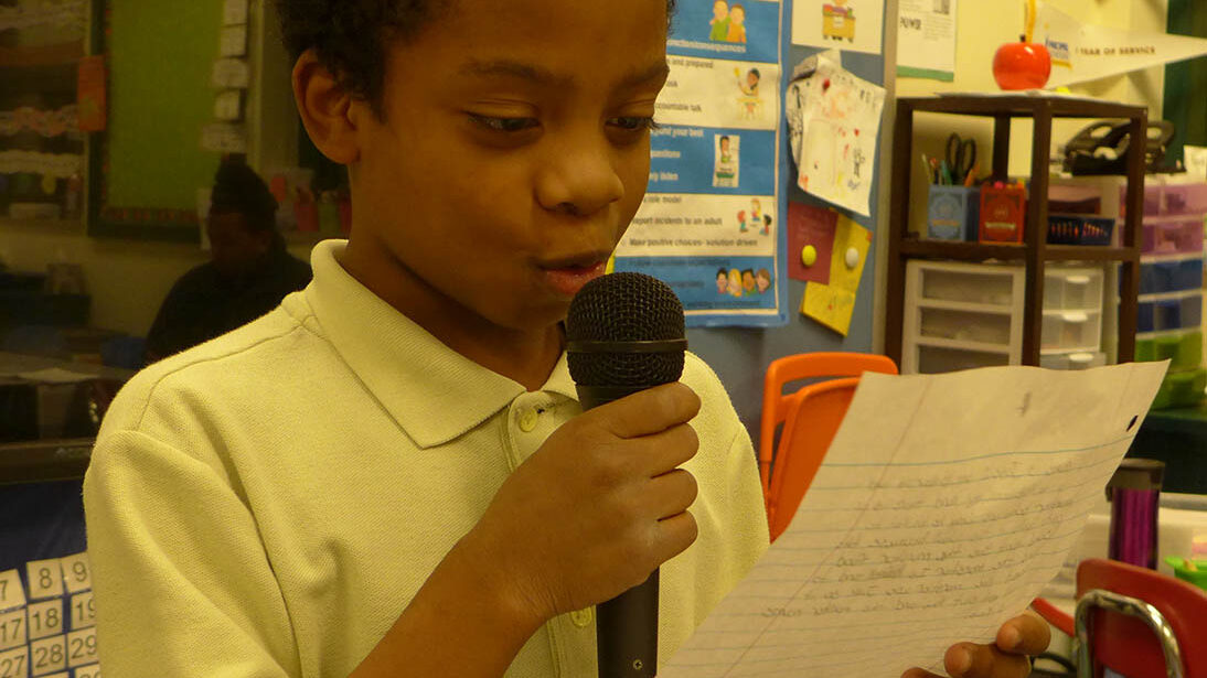 A youth holding a microphone reading a piece of paper in a classroom