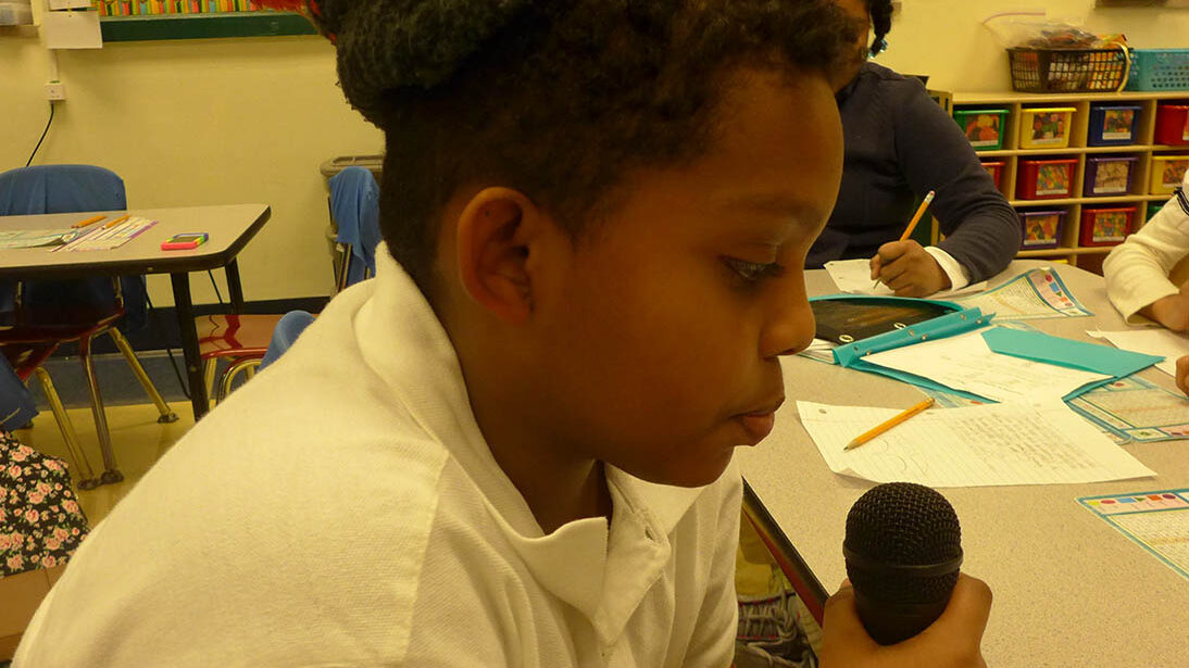 A youth holding and speaking into a microphone in a classroom