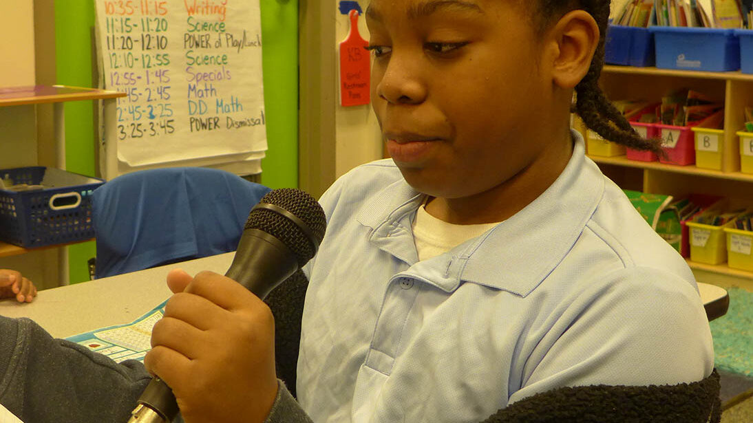 A youth holding and speaking into a microphone in a classroom