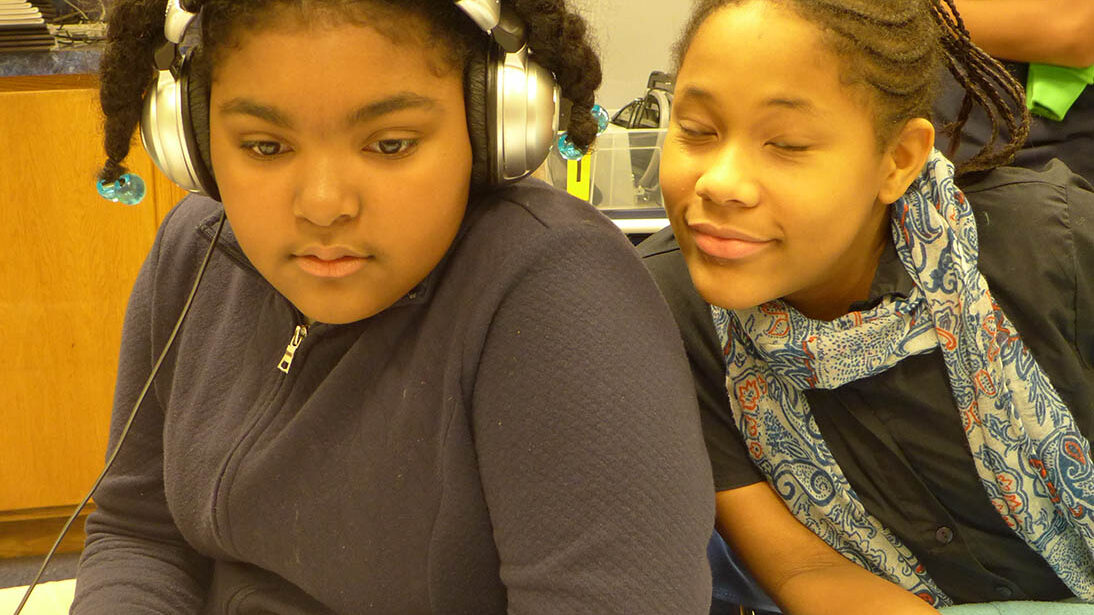 Two youth sitting in a classroom, one wearing headphones