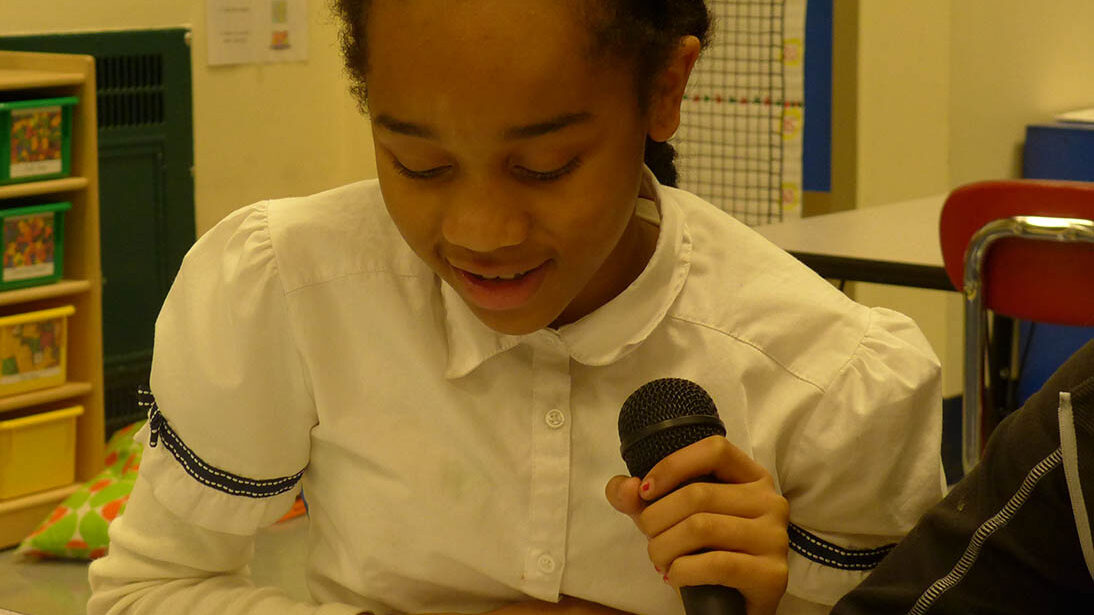 A youth holding and speaking into a microphone looking down at a piece of paper in a classroom