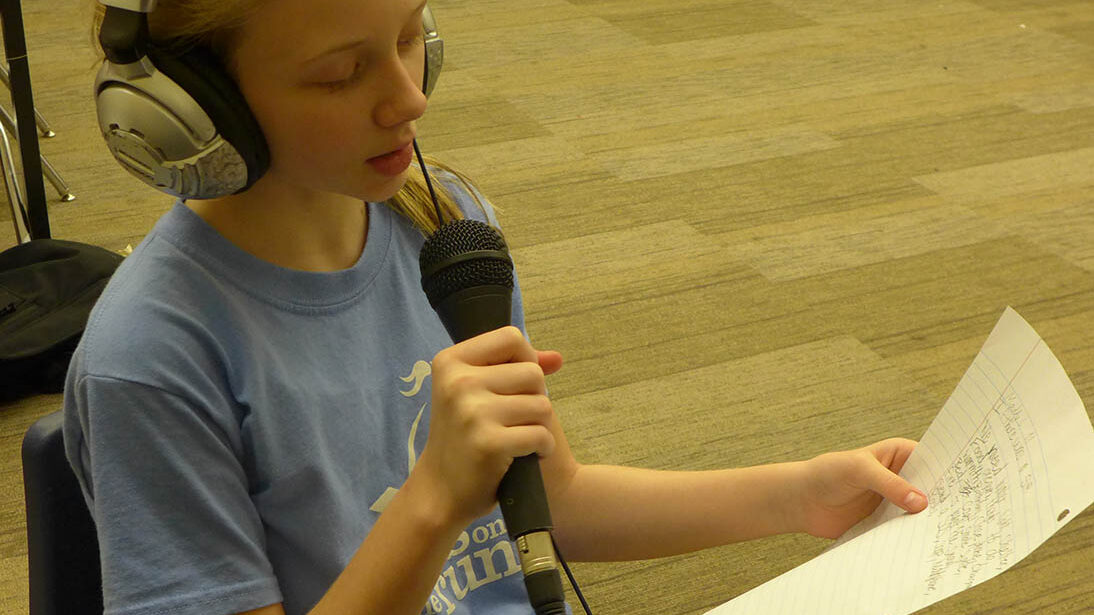 A youth wearing headphones reading a piece of paper into a microphone in a classroom