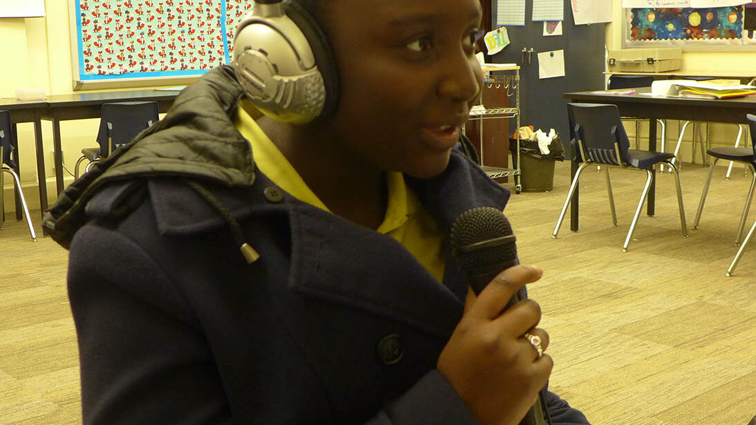 A youth wearing headphones holding and speaking into a microphone in a classroom