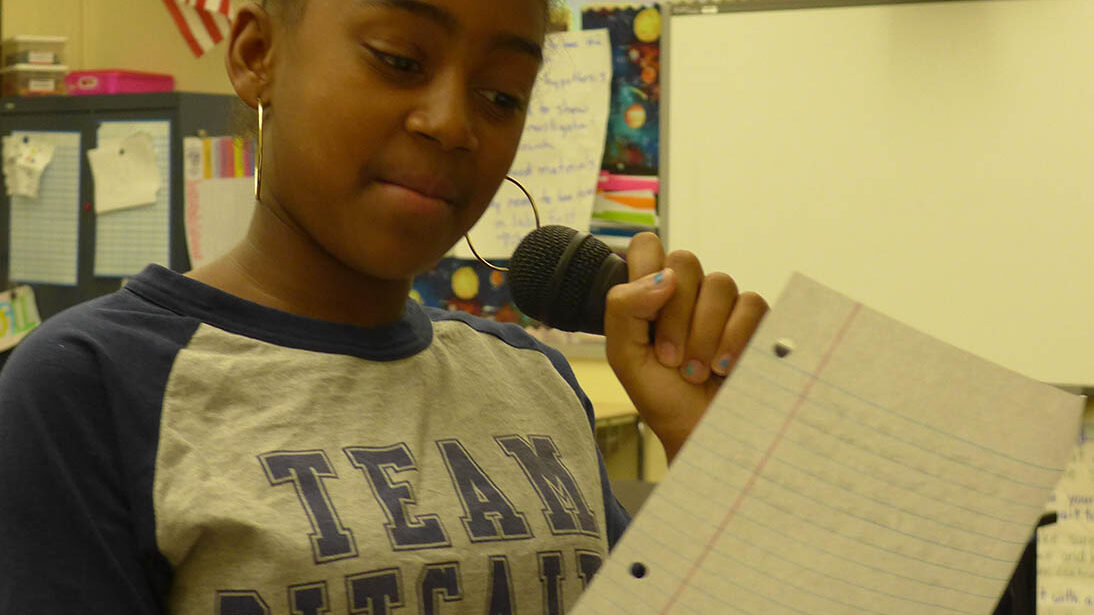 A youth holding a piece of paper reading into a microphone in a classroom