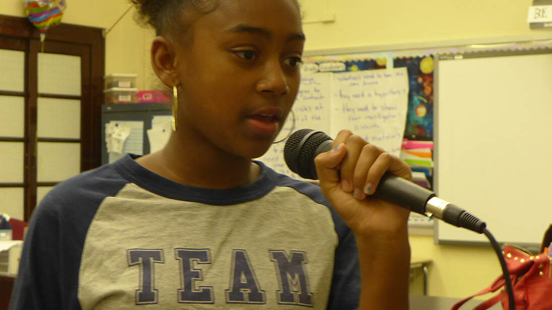 A youth holding and speaking into a microphone in a classroom