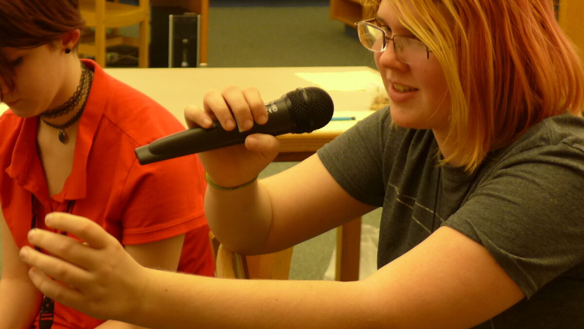Two teens sitting in a library, one holding and speaking into a microphone