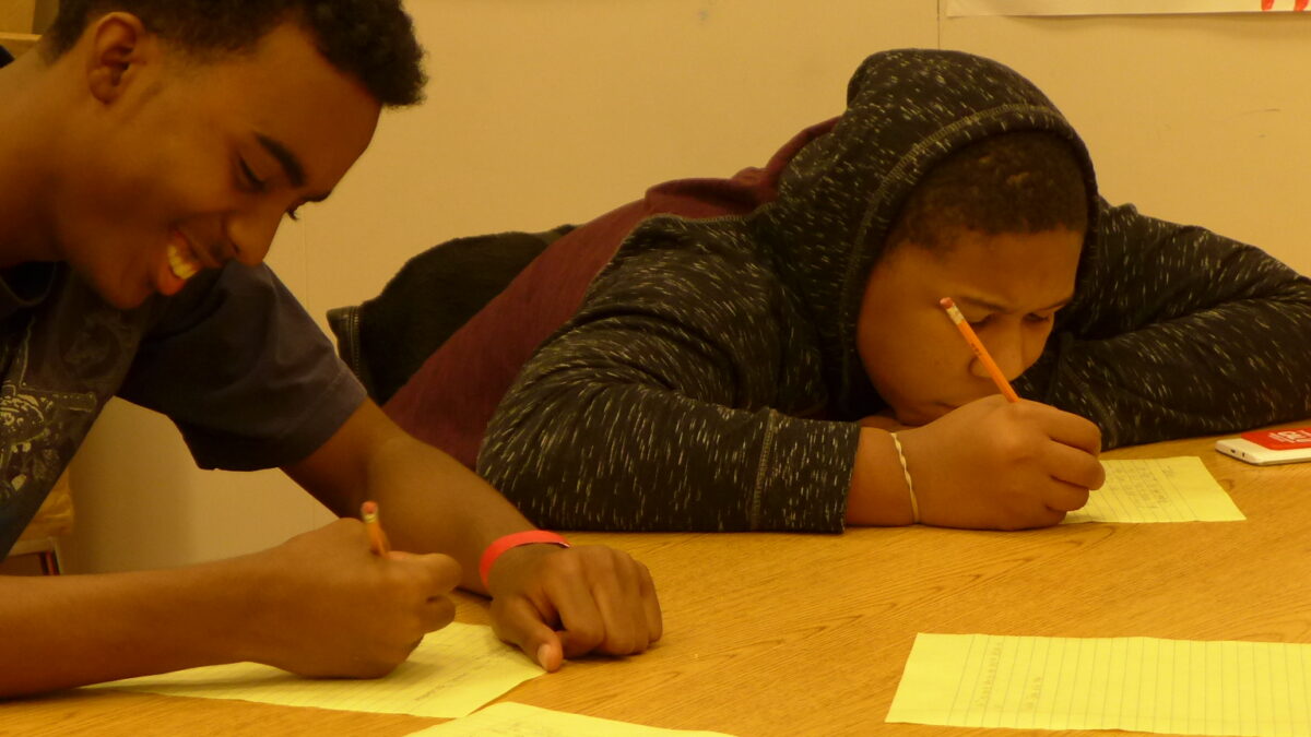 Two teens writing on pieces of paper on a table