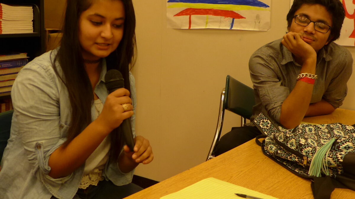 Two teens sitting at a table in a classroom, one holding a microphone