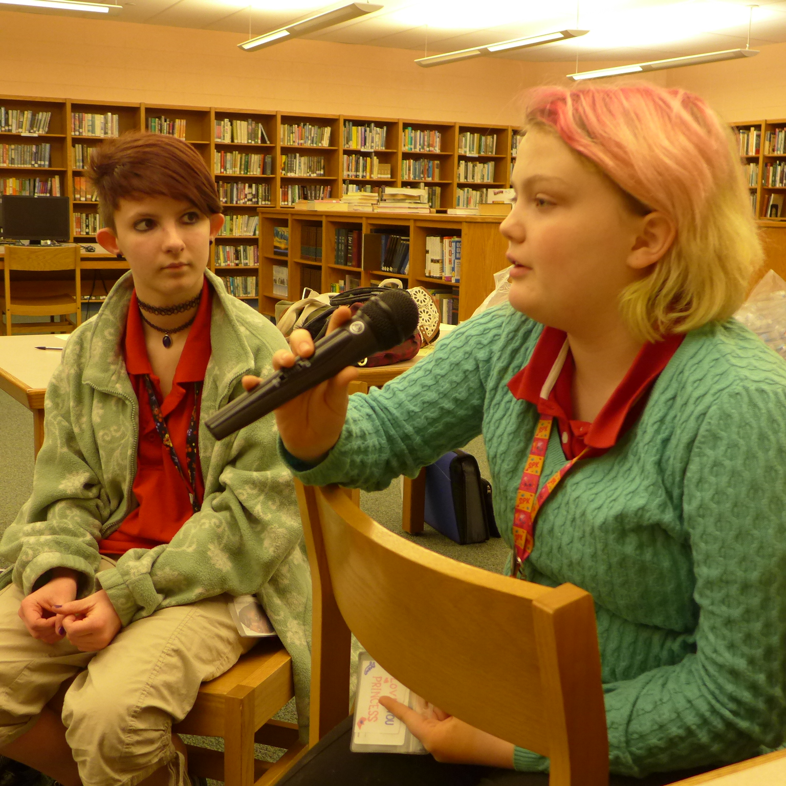 Two youth sitting in a library, one holding and speaking into a microphone