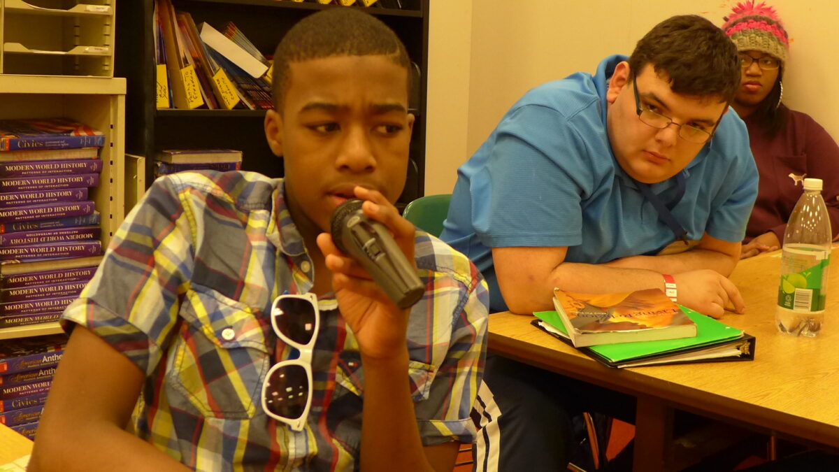 Three teens sitting in a classroom, one holding a microphone