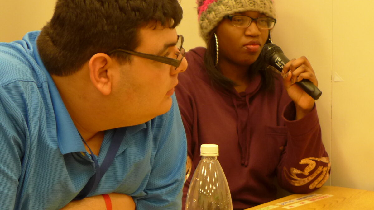 Two teens sitting in a classroom, one holding a microphone