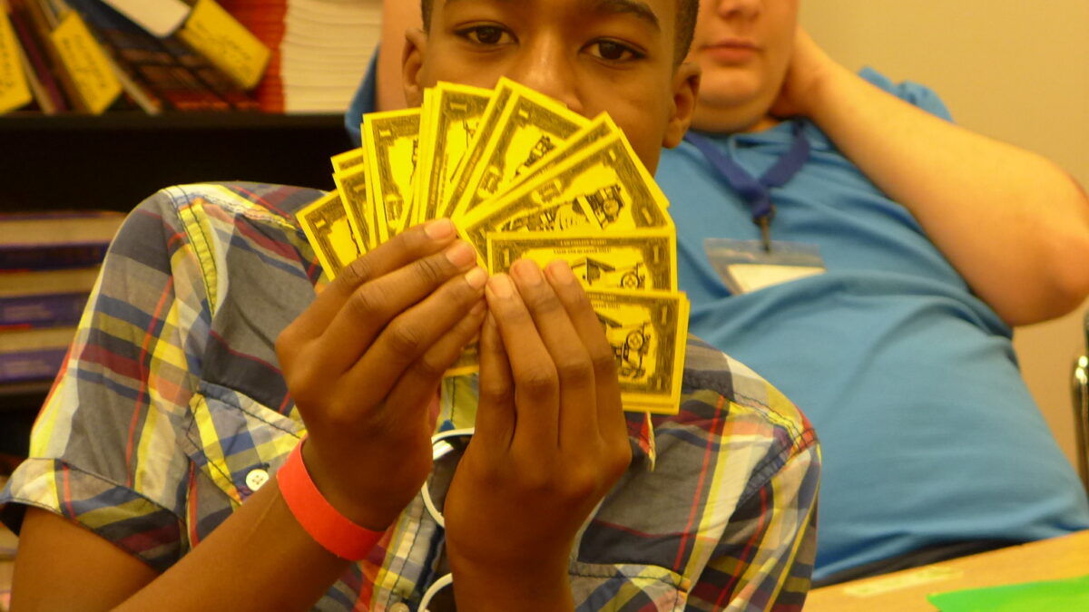 Two teens sitting at a table in a classroom, one holding a fan of board game bills