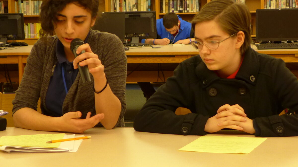 Two teens looking at manila folders on a table in a library, one holding and speaking into a microphone