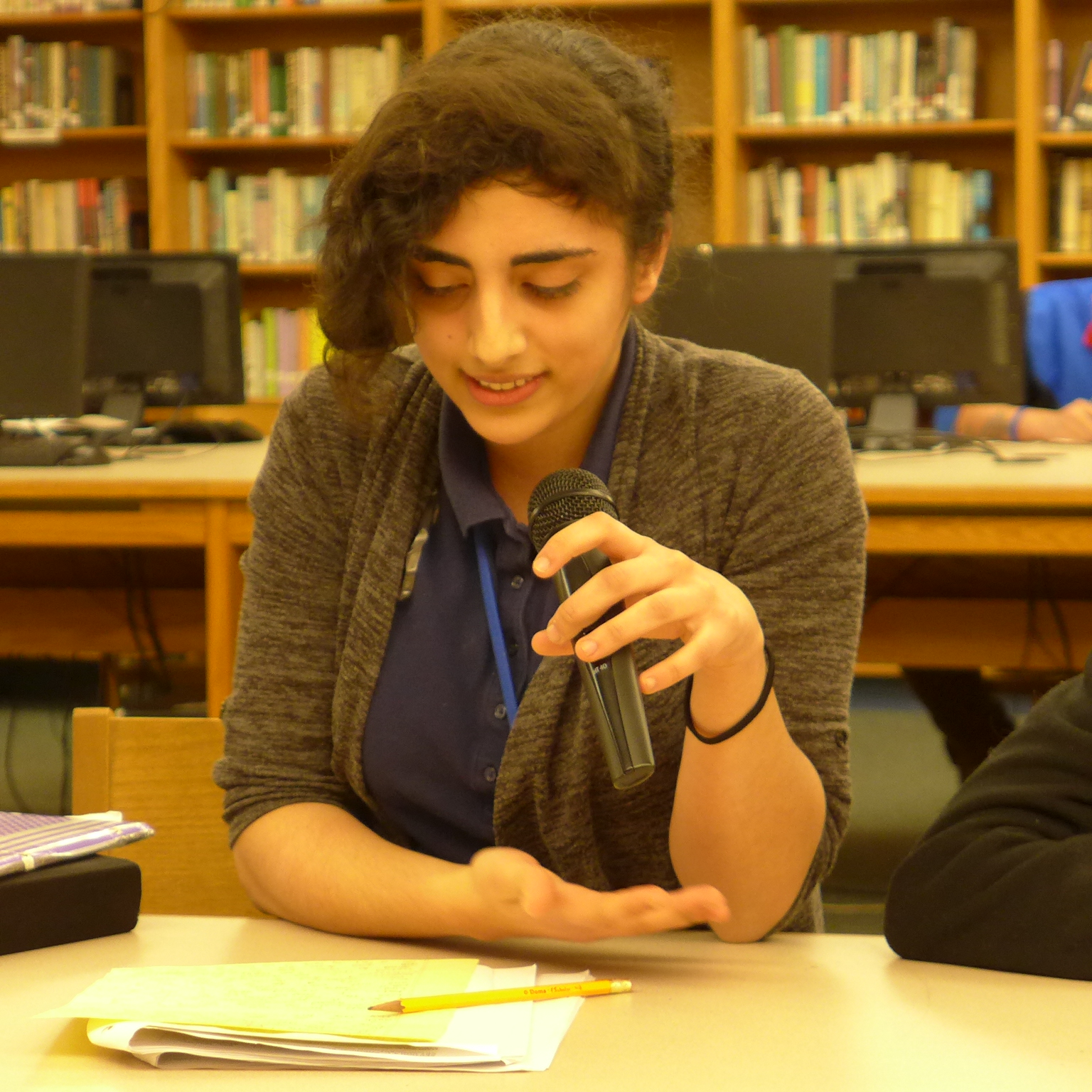 A teen looking down at a manila folder and holding and speaking into a microphone in a library