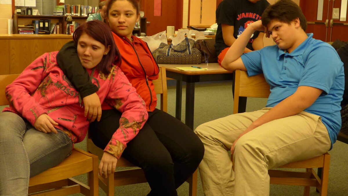 Three teens sitting in chairs and one teen sitting on a desk in a library