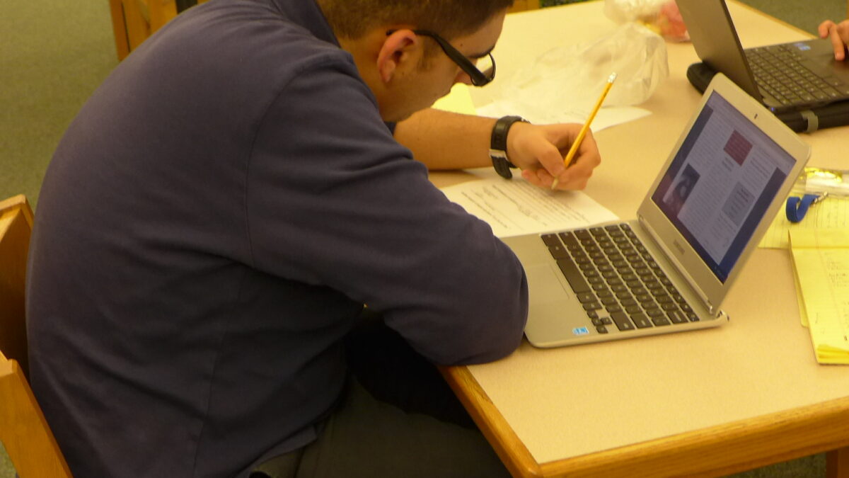 A youth sitting behind a laptop computer writing on a piece of paper
