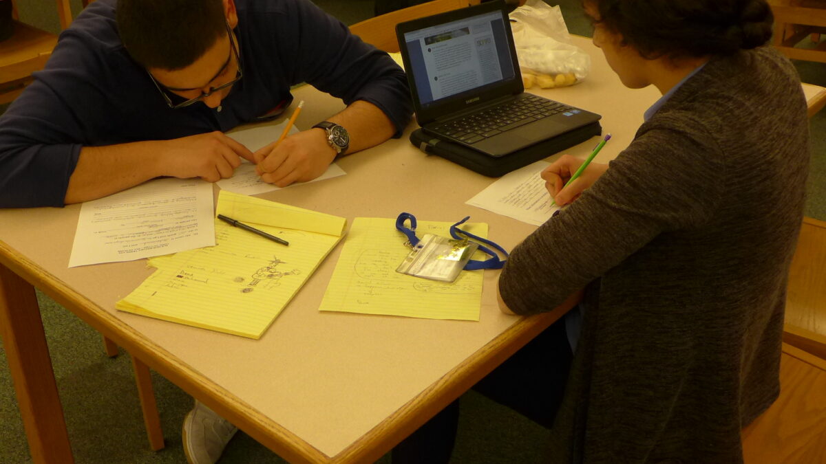 Two youth writing on pieces of paper on a table in a library