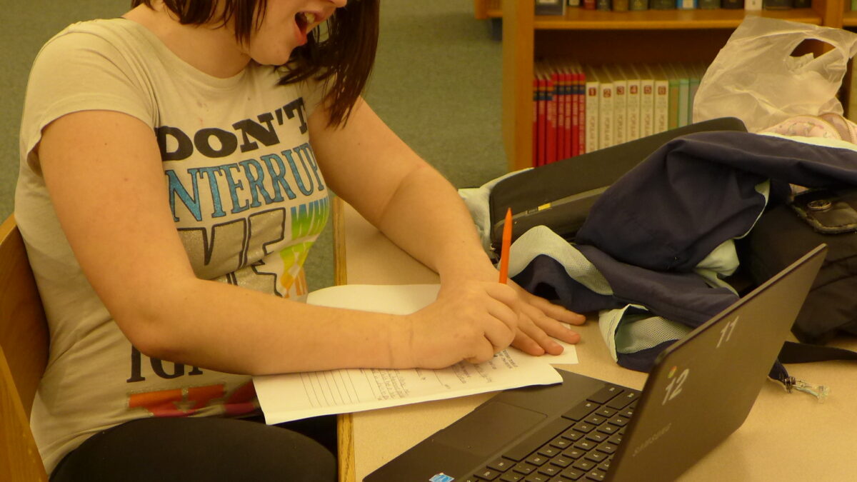 A youth sitting behind a laptop computer writing on a piece of paper