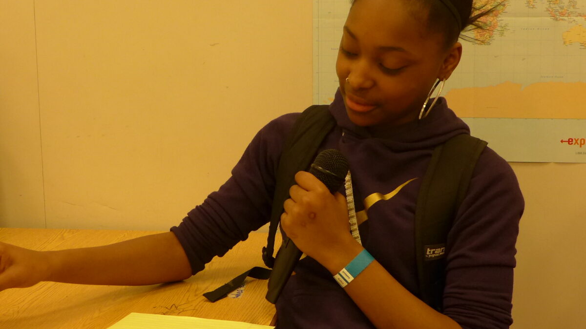 A teen holding a microphone looking down at a piece of paper in a classroom