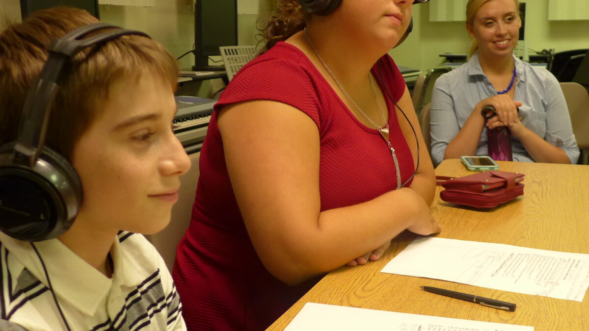 A youth and an adult wearing headphones sitting behind pieces of paper on a long wooden table