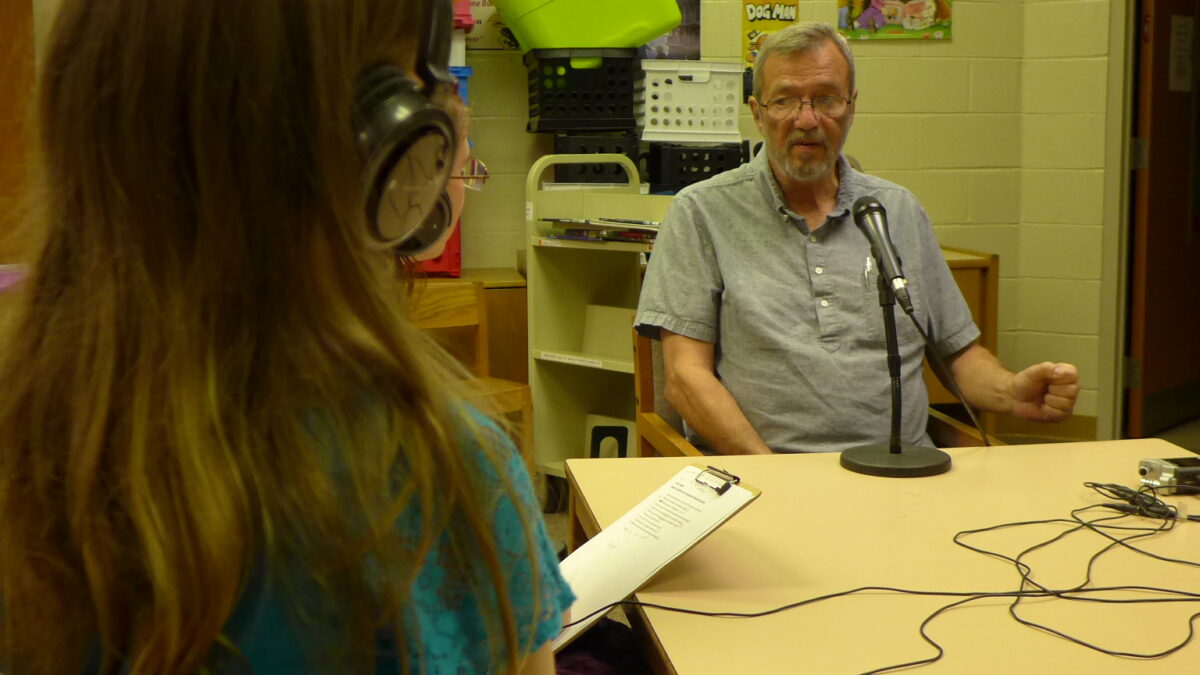 A youth wearing headphones interviewing an adult speaking into a microphone in a classroom