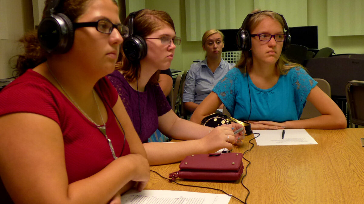 Three youth wearing headphones sitting behind pieces of paper on a long wooden table