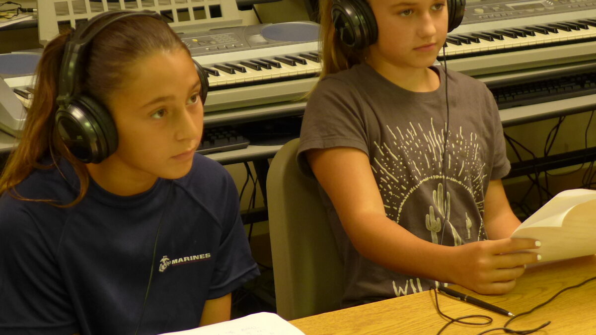 Two youth wearing headphones looking at pieces of paper on a long wooden table