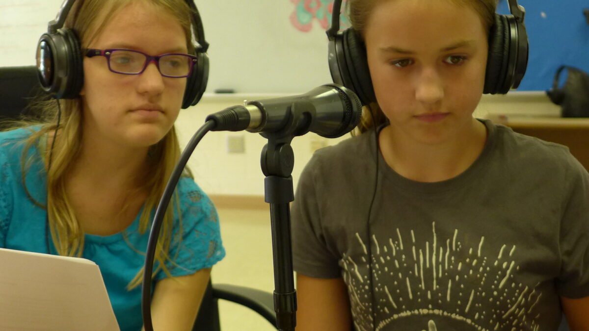 Two youth wearing headphones holding pieces of paper sitting behind a microphone in a classroom