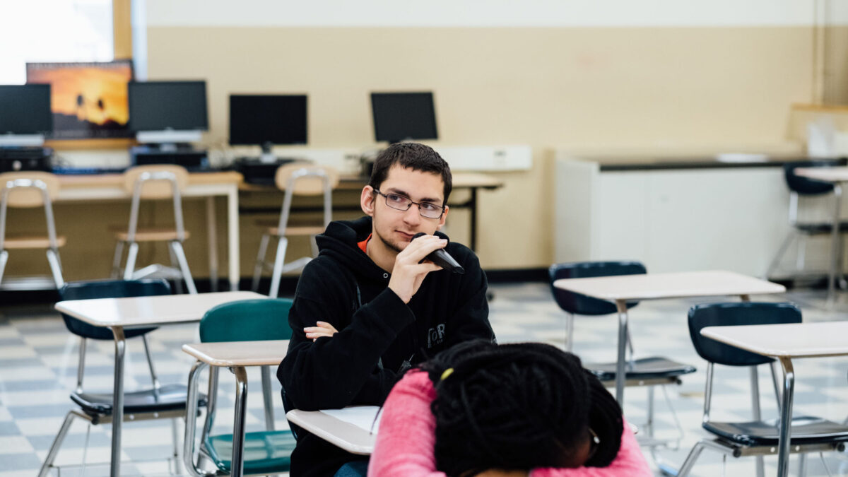 A teen speaking into a microphone and another teen sleeping in a classroom