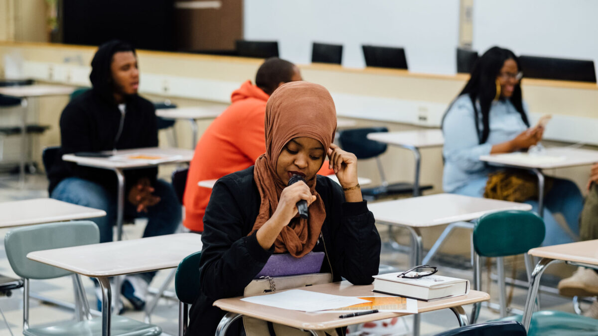Four teens sitting at classroom desks, one speaking into a microphone