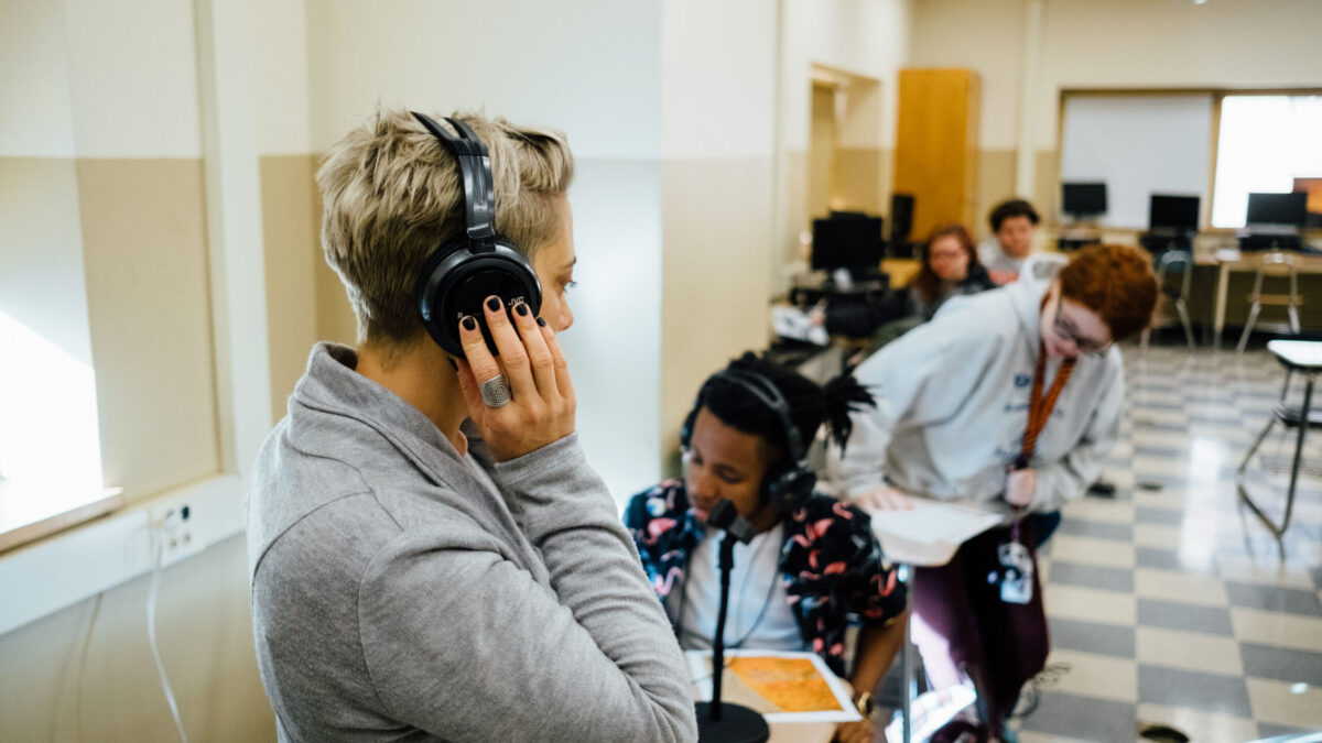 An adult wearing headphones and four teens sitting at classroom desks