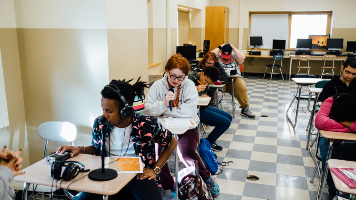 Seven teens sitting at classroom desks, one wearing headphones interacting with a portable recorder, one speaking into a microphone