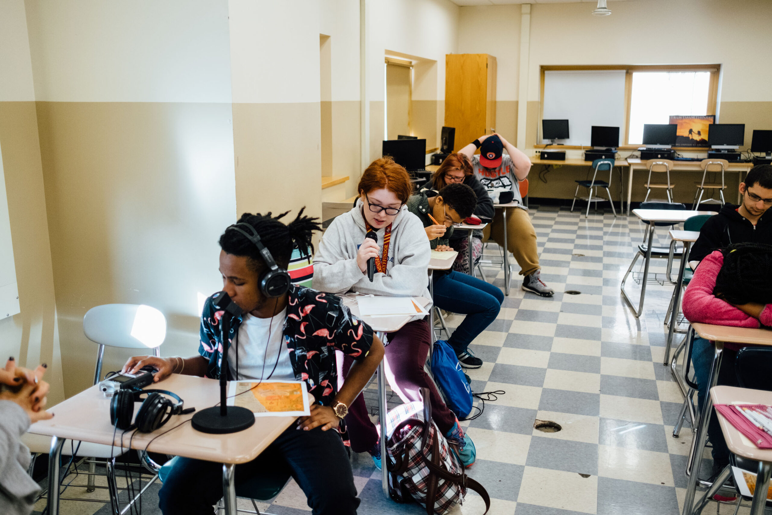 Seven teens sitting at classroom desks, one wearing headphones interacting with a portable recorder, one speaking into a microphone