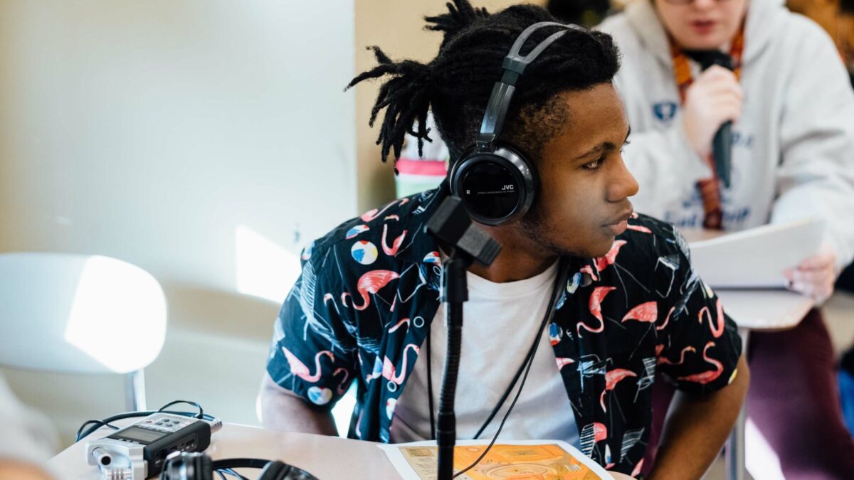 A teen wearing headphones sitting behind a microphone on a classroom desk and a teen speaking into a microphone sitting at a classroom desk