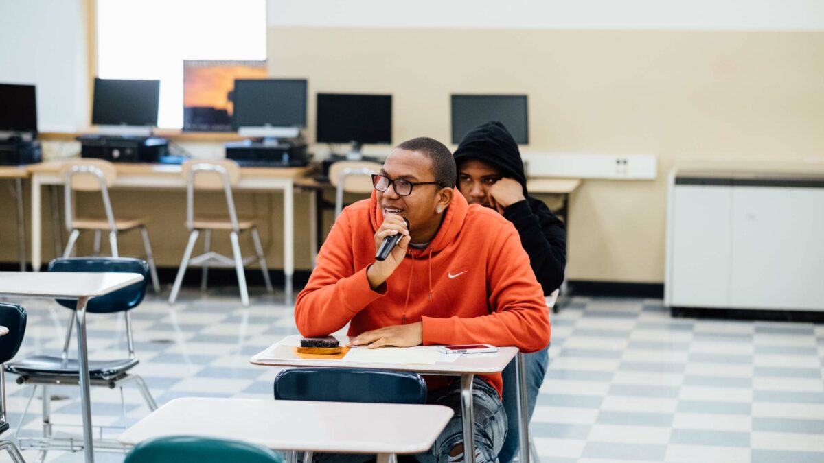 A teen speaking into a microphone sitting at a desk in a classroom
