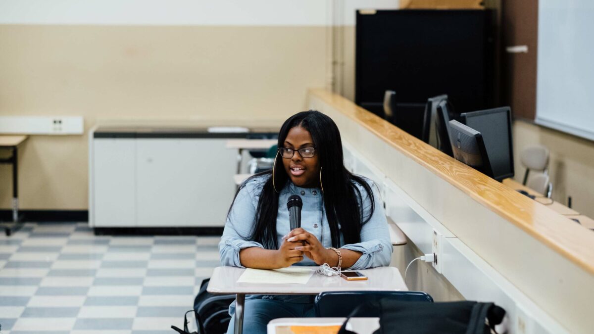 A teen speaking into a microphone sitting at a desk in a classroom
