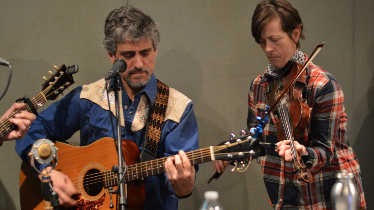 An adult playing a fiddle and an adult playing an acoustic guitar in a radio studio