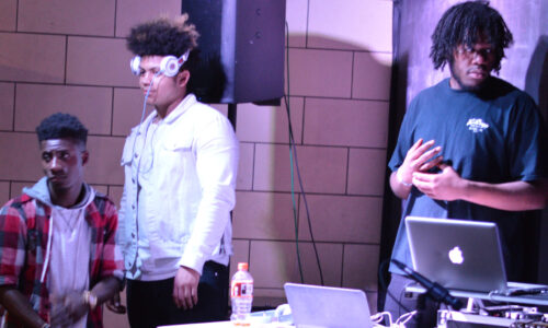 Three teens standing behind a laptop and a turntable in front of a speaker