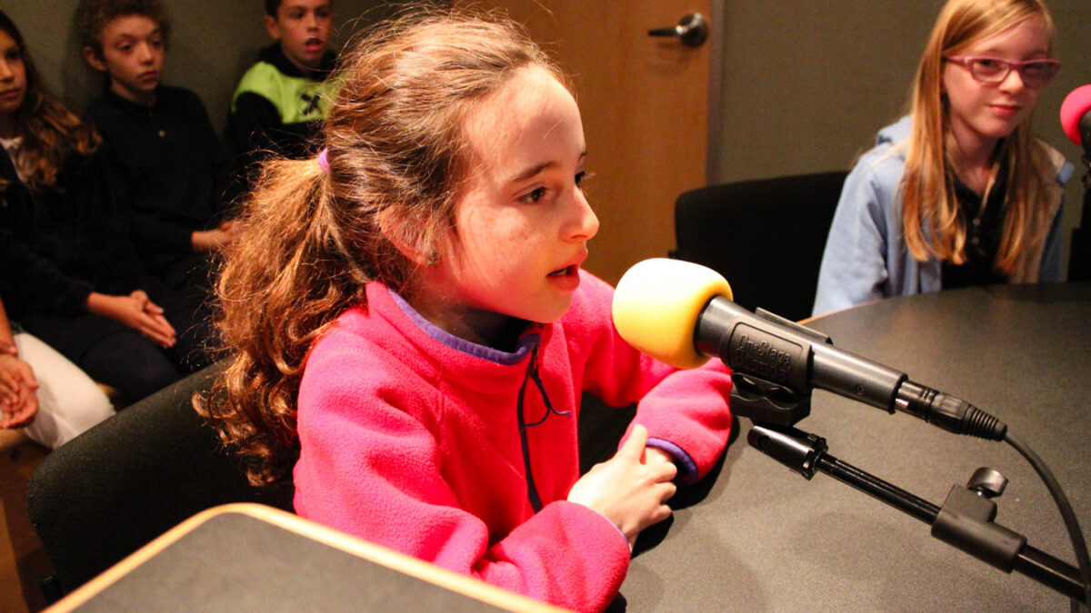 Two youth speaking into microphones as two youth look on in the background in a radio studio