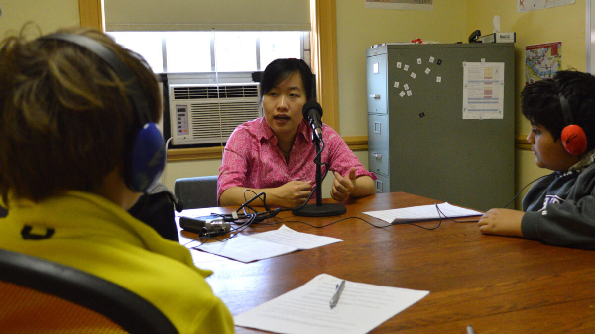 Two youth wearing headphones interviewing an adult speaking into a microphone in a library
