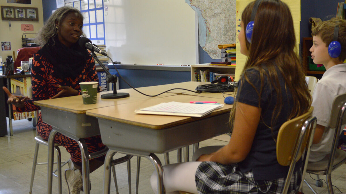 Two youth wearing headphones interviewing an adult speaking into a microphone in a classroom
